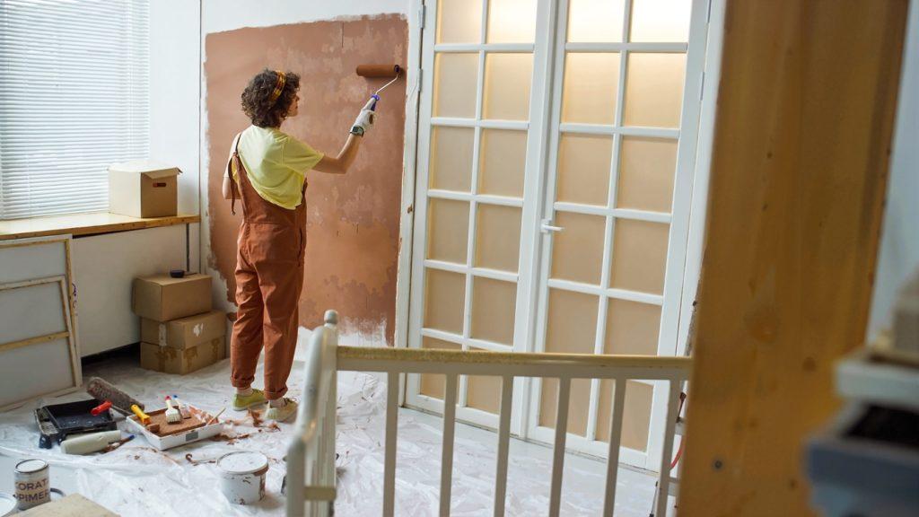 woman painting wall with roller brush in room filled with painting supplies