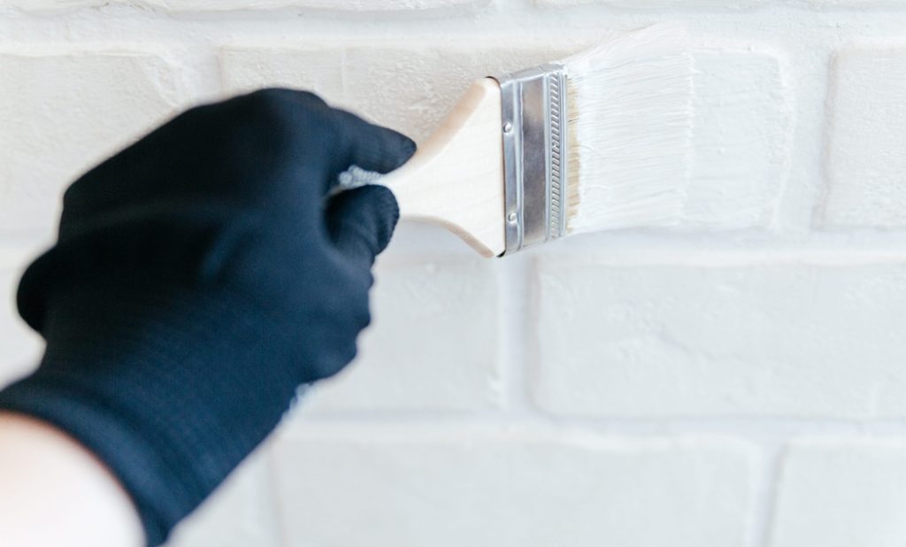 workers hand with a brush painting a brick wall with white paint