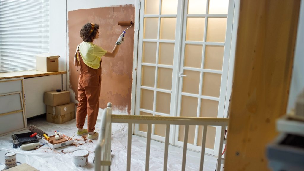 woman painting wall with roller brush in room filled with painting supplies